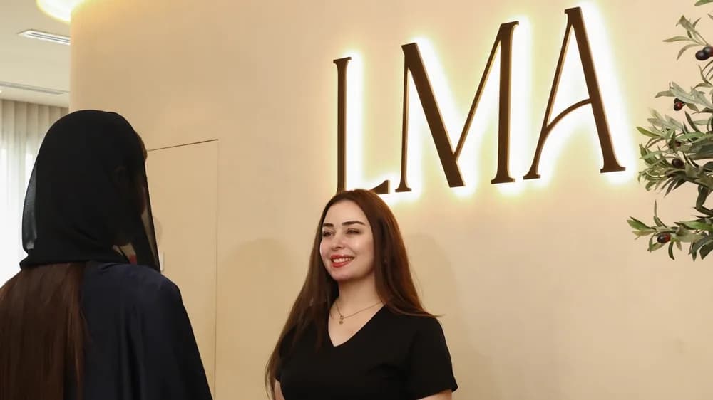 A receptionist in black greets a client in a black abaya and shayla under the backlit LMA sign at LMA Clinic in Dubai