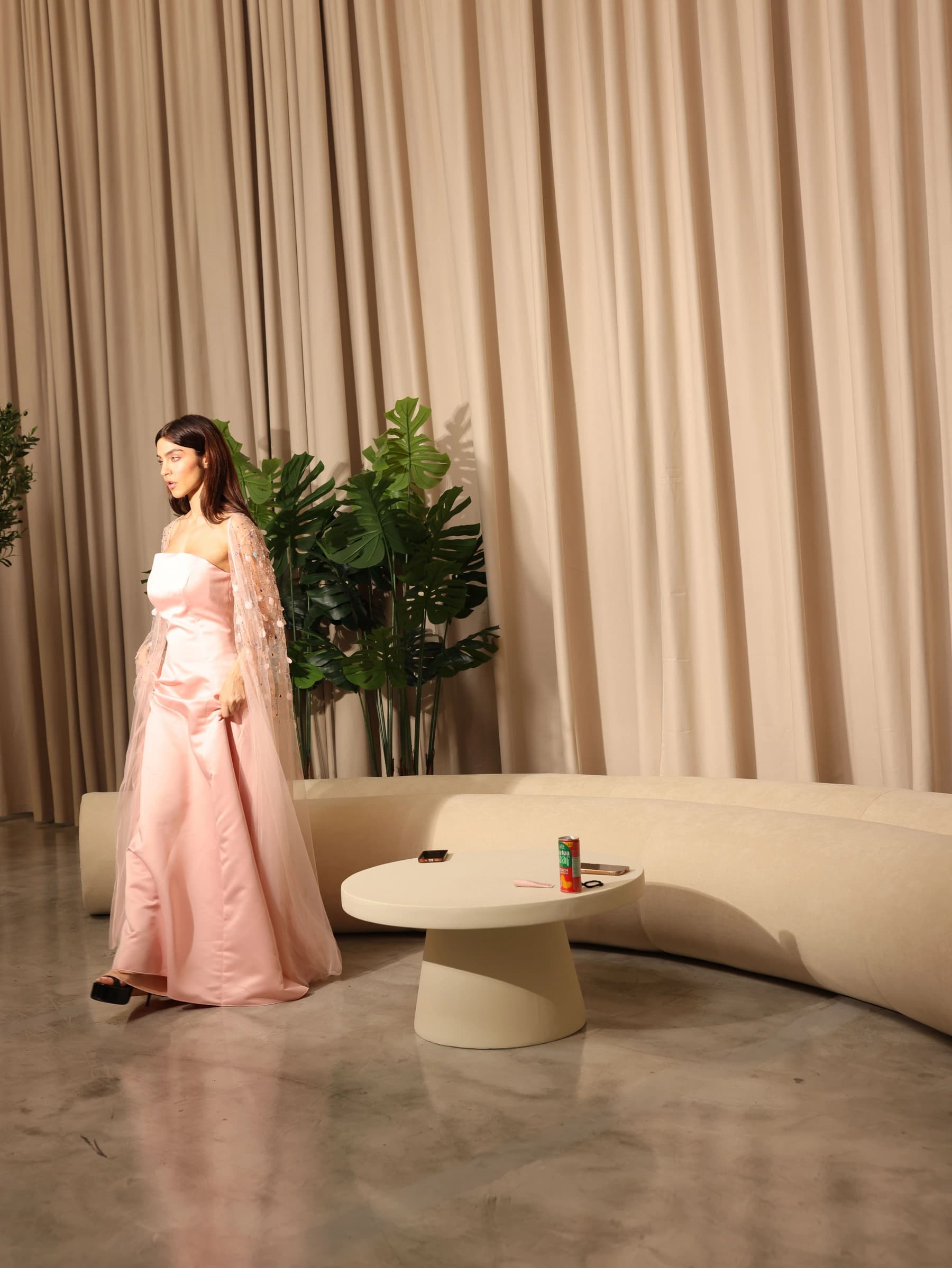 Model in a pink satin gown and sequined cape walking past the curved sofa in the Lobby room during a shoot