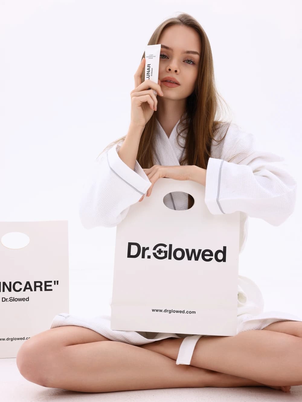 عارضة بروب أبيض تحمل كريم Lunar المجدِّد من Dr.Glowed خلف كيس ورقي يحمل اسم العلامة