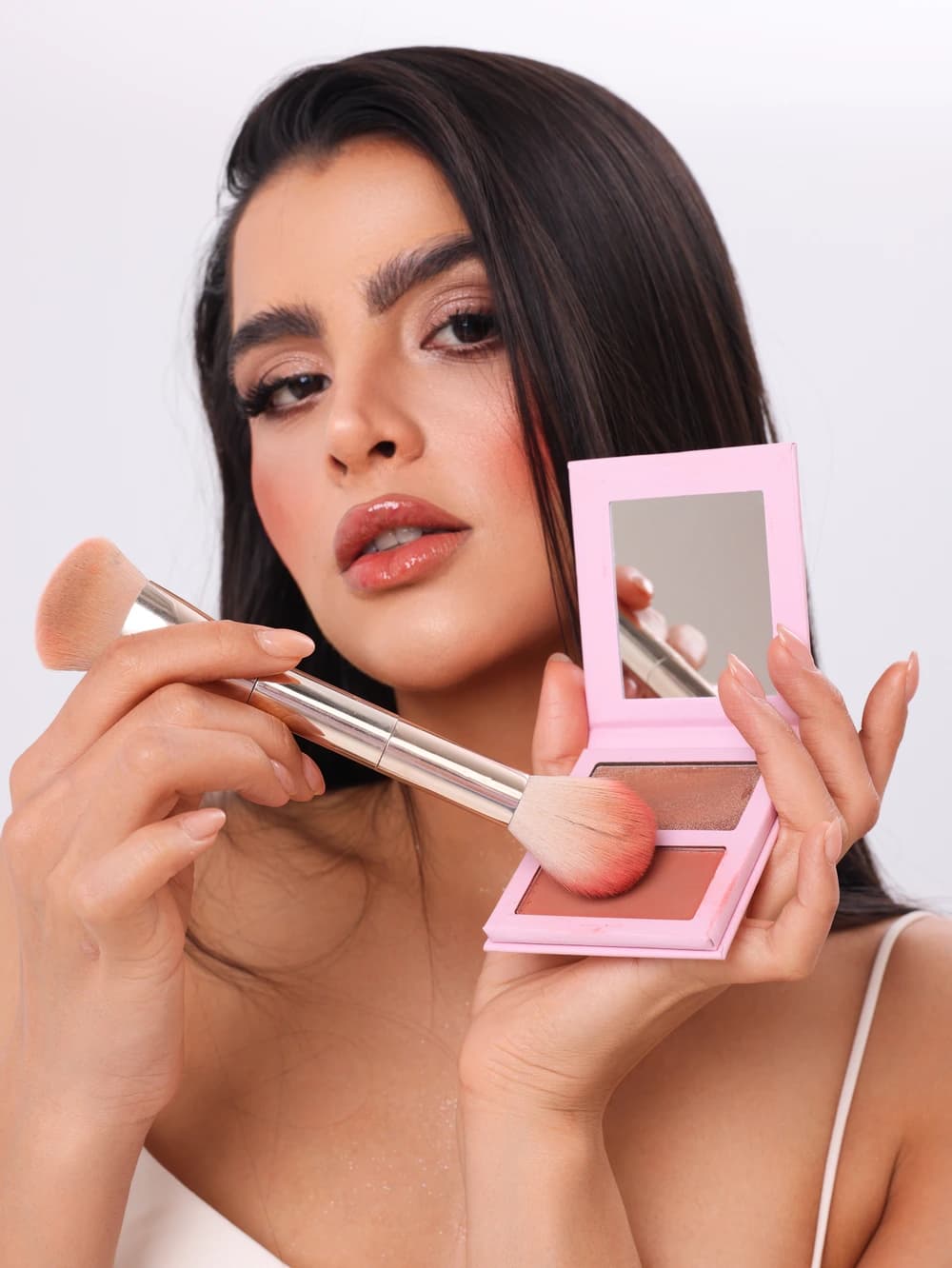 A model holding a pink blush palette and a brush to her cheek