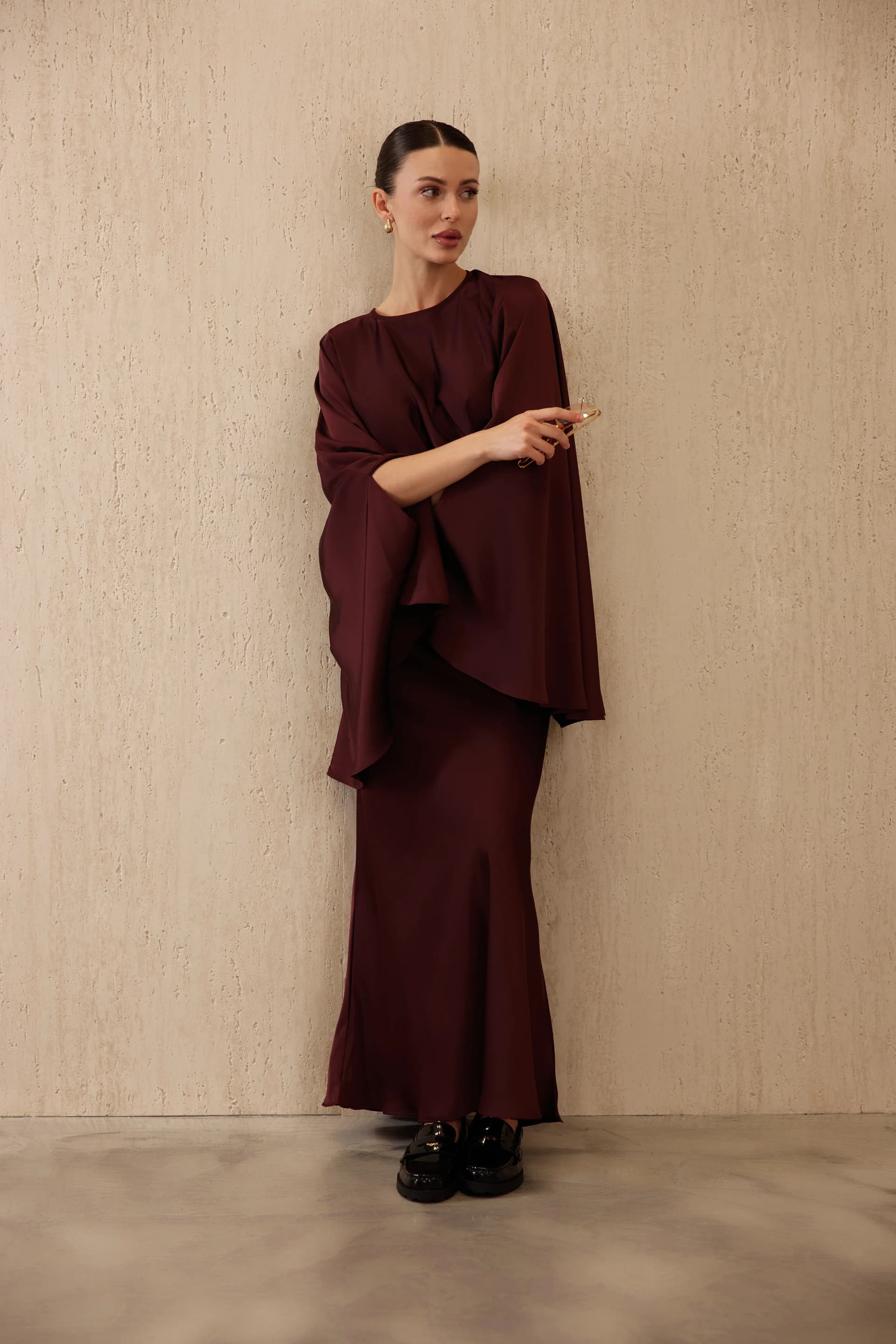 Model in a long burgundy dress with wide cape sleeves against a textured plaster wall