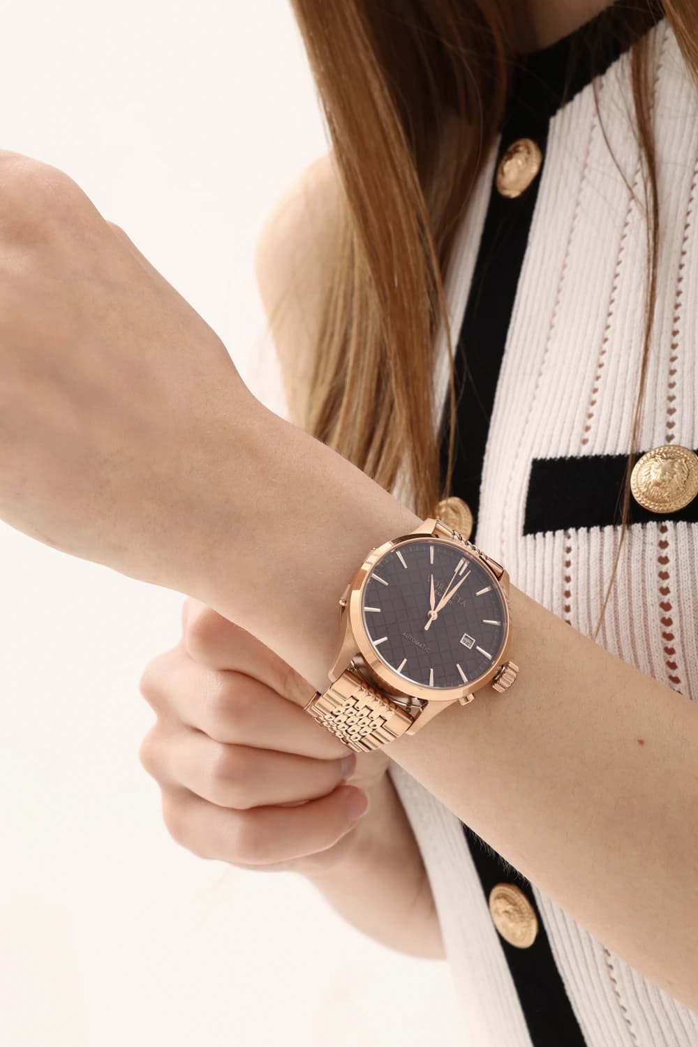 A rose-gold automatic watch on a model's wrist, over a cream knit with gold buttons