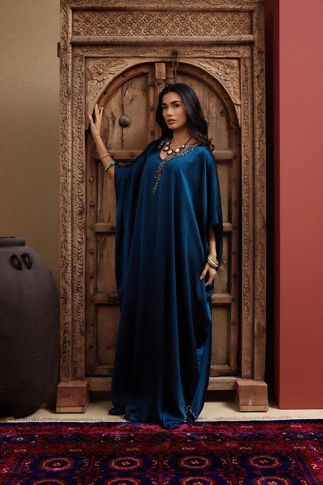 Model in a deep teal satin kaftan with a beaded neckline, one hand on a carved wooden door in the Heritage room, a patterned rug at her feet